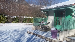 Un espace extérieur aménagé avec des tables et des chaises, recouvert de neige, un petit bâtiment avec une porte verte et une poubelle violette.