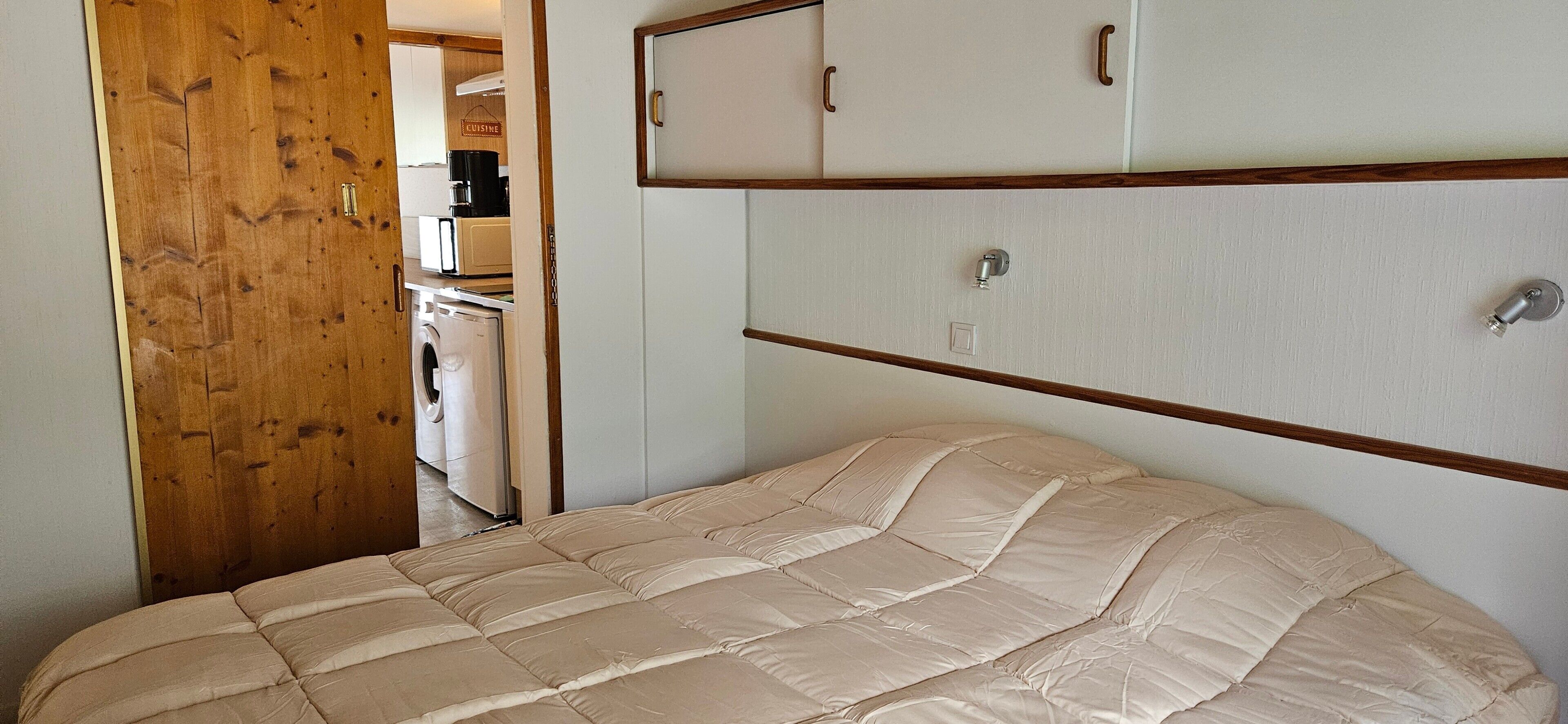 Quarto com cama, guarda-roupa de madeira e uma kitchenette ao fundo.