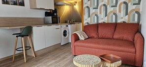 Un espace de vie compact comprenant un canapé, une table basse ronde en bois, un lave-linge, un four à micro-ondes et une chaise.