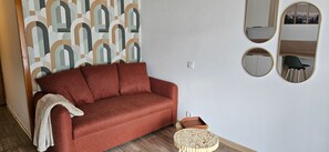 Un canapé rouge avec un plaid blanc, un tabouret rond en bois et un mur recouvert d’un papier peint à motifs géométriques, ainsi que trois miroirs.