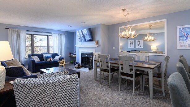 A living room with a fireplace, a dining area with a chandelier, and a TV mounted on the wall. - Ski-In/Out Winterplace Okemo Mountain Condo-Bldg M (Ludlow)