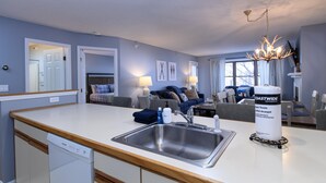 A modern kitchen with a built-in dishwasher, a stainless steel sink, and a large island. There is a dining area with a table and chairs, and a living room with a sofa and a fireplace. - Ski-In/Out Winterplace Okemo Mountain Condo-Bldg M (Ludlow)