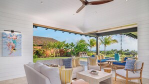 Un soggiorno moderno con un divano bianco, cuscini a righe e un tavolino da caffè. Una grande finestra offre vista su una piscina e piante tropicali.