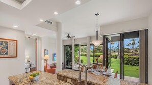 A modern kitchen with a granite countertop, a sink, and a dining area with a view of a lush garden. - Mauna Lani Point J101 - Enhanced Comfort & Cleanliness | Professionally Managed (Kamuela)