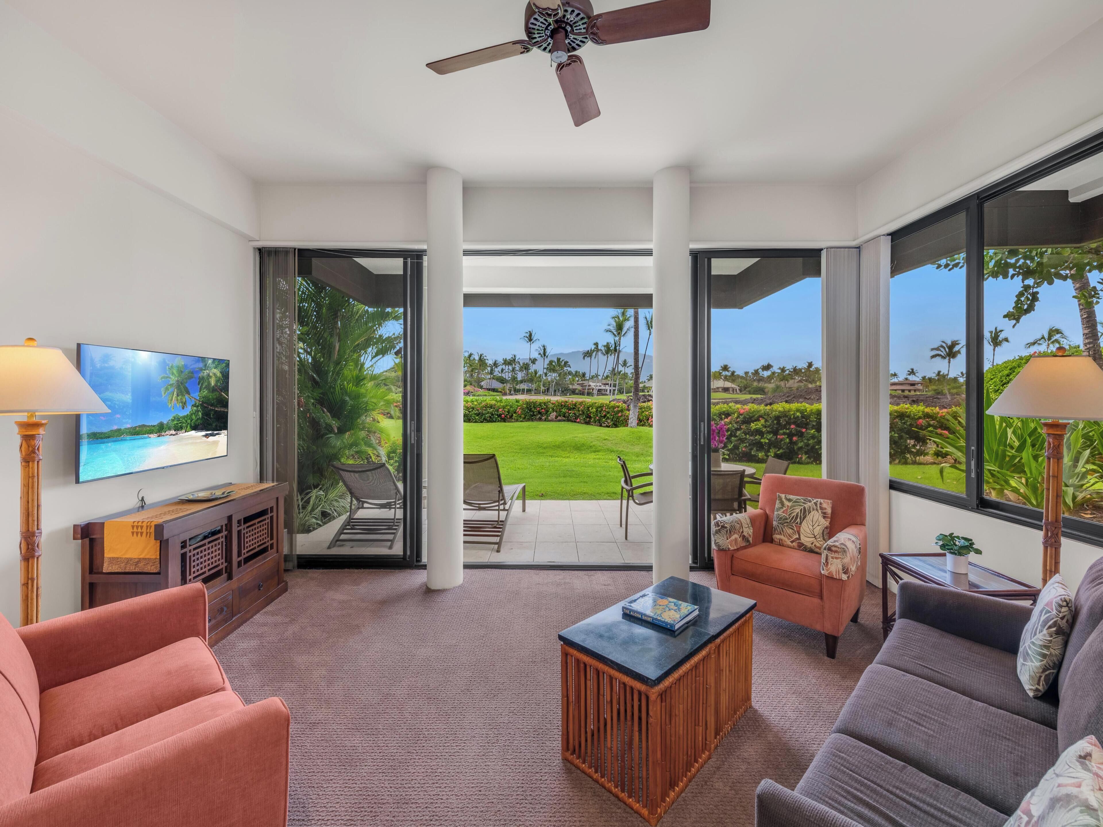 A spacious living room with a large TV, comfortable seating, a ceiling fan, and a view of a lush garden and beach.