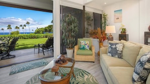 A spacious living room with a view of the ocean, a large window, and a tropical-themed decor. - *CoralTree Managed* Ground Floor Ocean & Golf Views - Mauna Lani Point A107 (Kamuela)
