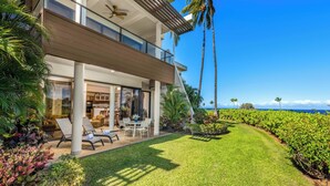 A modern two-story house with a balcony, a patio with outdoor furniture, and a well-maintained lawn. - Mauna Lani Point B102 is a Golfer's Dream w/Fairway and Ocean Views | CoralTree (Kohala Coast)