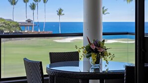 A dining area with a table and chairs, a vase with flowers, and a view of the ocean and palm trees.