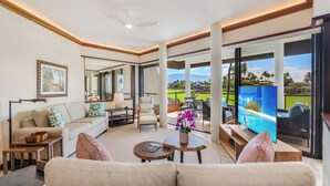 A spacious living room with a large sofa, a coffee table, and a TV. There is a balcony with a view of a golf course and mountains. - Modern & Renovated Mauna Lani Point F204 | Ocean Views | Professionally Managed (Kamuela)