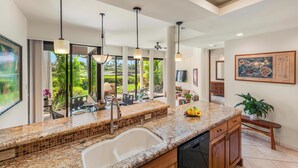 Cocina moderna con encimeras de granito, pileta doble y lavavajillas. Se observan luces colgantes, un amplio ventanal con vista a un jardín y un comedor visible a través del ventanal.