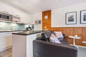 Une cuisine moderne équipée d’un four à micro-ondes, d’une bouilloire et d’un support pour la bouilloire. On y trouve un petit îlot central avec un évier, ainsi qu’un canapé en cuir noir agrémenté de coussins à motifs floraux. Une table ronde, sur laquelle sont posés une bougie et un tableau encadré, se trouve à côté du canapé.