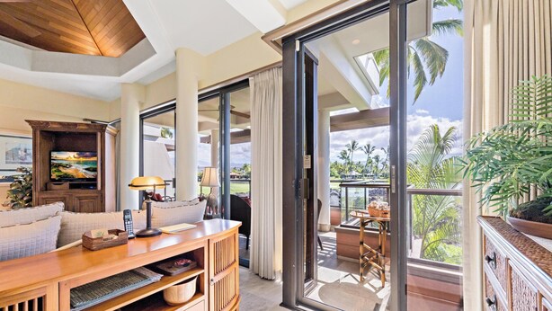 A spacious living area with a wooden entertainment unit, a comfortable sofa, and a large sliding glass door leading to a balcony with a view of palm trees and a clear sky. - Mauna Lani Point H207 - Includes Membership to Nearby Luxury Fitness Club! (Kamuela)
