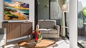 Ein modernes Wohnzimmer mit einem großen Gemälde von einem Strandsonnenuntergang, einem grauen Sofa, einem Holztisch mit Blumen in einer Vase und einem Glasgeländer.