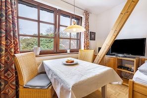 Une pièce comprenant une table à manger, des chaises en osier, un téléviseur et une fenêtre avec des rideaux.