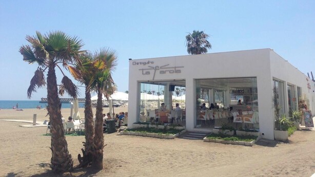 Un ristorante sulla spiaggia con una facciata bianca moderna, tavoli all'aperto e palme.