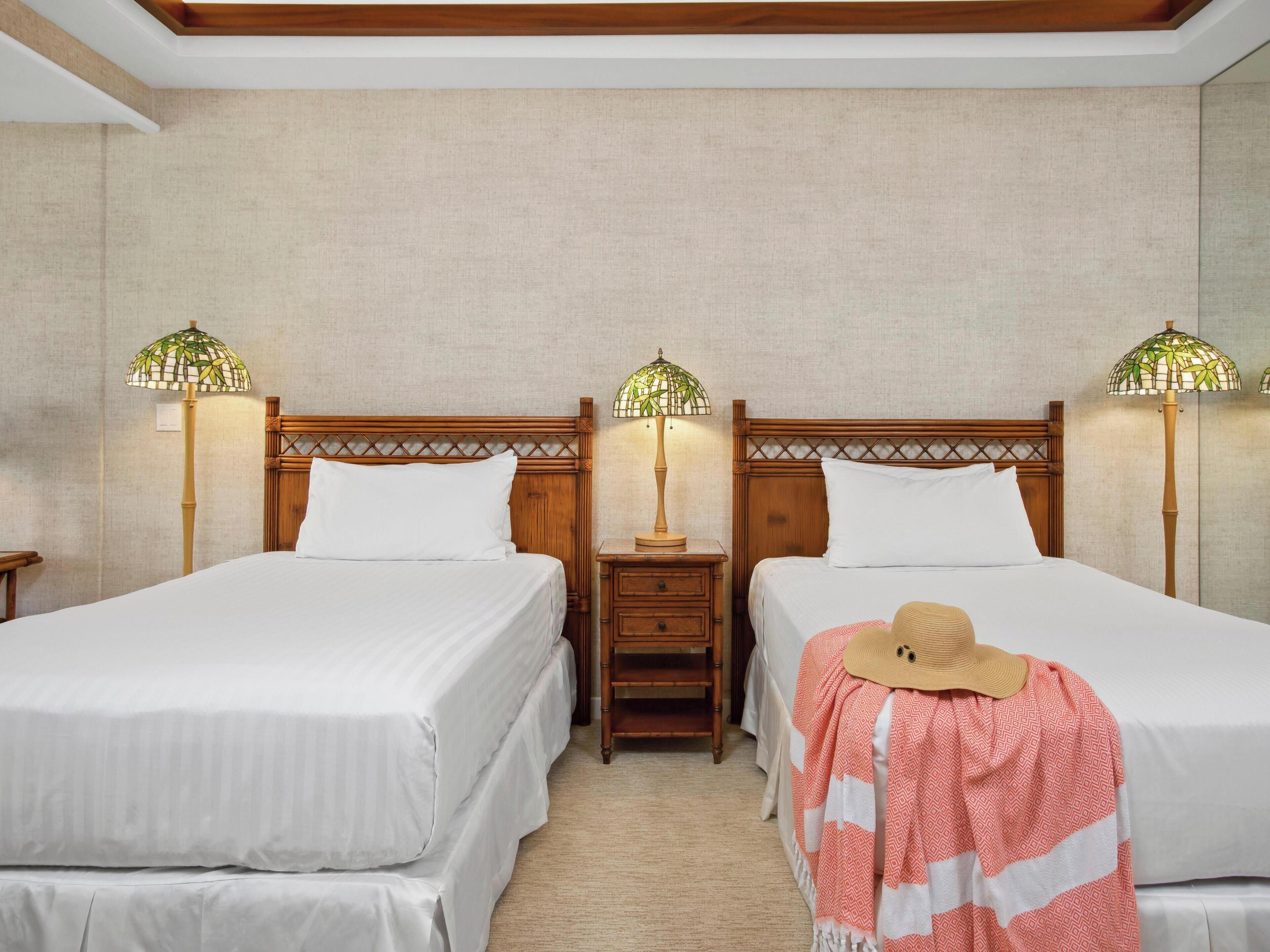 Habitación de hotel con dos camas individuales, cabeceros de madera, mesitas de noche con lámparas y alfombra en tonos neutros.