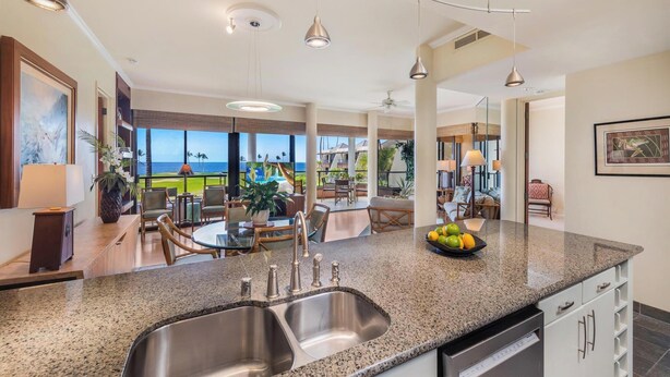 Una cocina moderna con encimera de granito, lavaplatos de acero inoxidable y muebles blancos. Hay una zona de comedor con mesa y sillas, y vistas al océano a través de amplias ventanas.