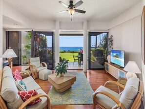 A spacious living room with a large TV, comfortable seating, and a view of the ocean through sliding glass doors.