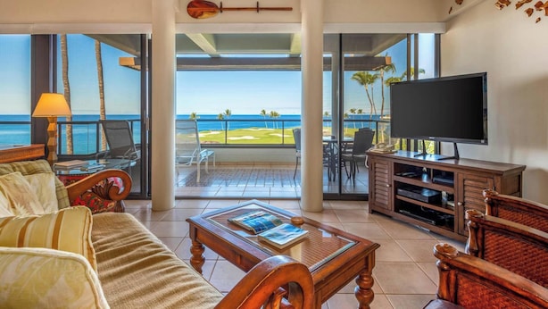 A spacious living room with a large TV, comfortable seating, a coffee table, and a view of the ocean.