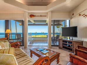 A spacious living room with a large TV, comfortable seating, a coffee table, and a view of the ocean.