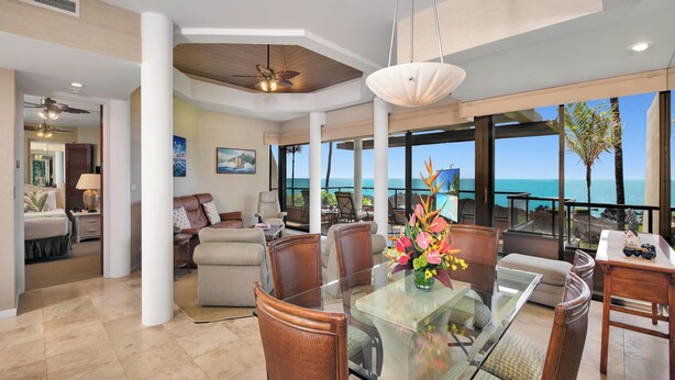 A spacious living area with a glass dining table, comfortable seating, and a view of the ocean. - Discounted Golf Rates! Mauna Lani Point E203 w/Ocean Views - CoralTree Managed (Kamuela)