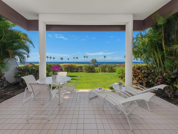 A covered patio with a view of the ocean, white chairs, and a table with a vase of flowers.