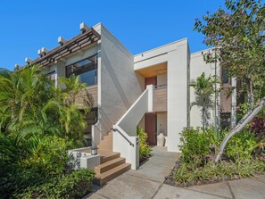 A modern two-story house with a staircase leading to the entrance, surrounded by lush greenery and a clear blue sky.