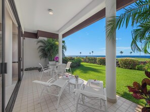 A covered outdoor seating area with white chairs and tables, overlooking a well-maintained lawn and the ocean.
