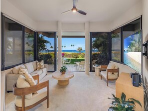 A spacious living room with a large window offering a view of the ocean and a balcony with outdoor furniture. - Golfer's Dream w/Fairway and Ocean Views | Mauna Lani Point B104 by CoralTree 🌊🌴 (Kamuela)