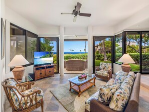 A spacious living room with a large flat-screen TV, wicker furniture, and a view of the ocean through large glass doors. - 🌊🌴😎 Ground Floor & Great for Families! Ocean View Mauna Lani Point C101 | Pool (Kamuela)