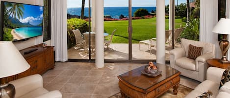 A living room with a large TV, comfortable seating, a coffee table, and a view of the ocean.