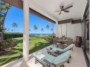 A covered outdoor area with a view of the ocean, a patio with lounge chairs, a table, and a ceiling fan. - Mauna Lani Point E106 | Sunset Views | Pool & Hot Tub | Sauna - CoralTree (Kamuela)