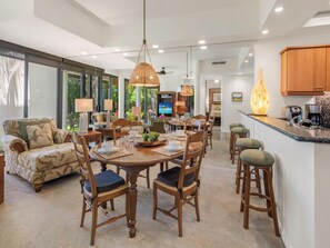 A spacious living area with a dining table, chairs, a sofa, and a kitchen area with a coffee machine. - Mauna Lani Point E106 | Sunset Views | Pool & Hot Tub | Sauna - CoralTree (Kamuela)