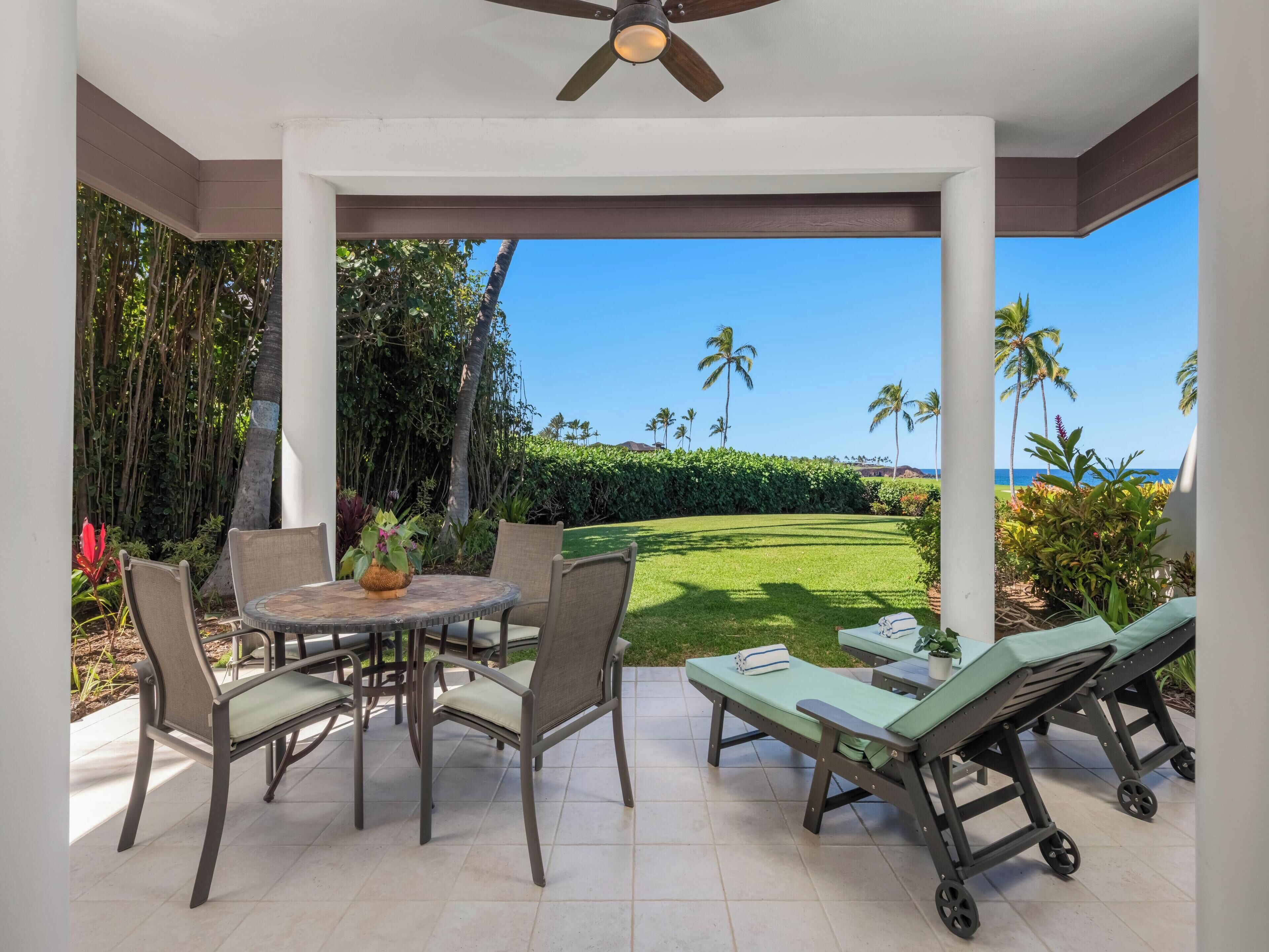 Une terrasse couverte avec une table et des chaises, un ventilateur de plafond et une vue sur un espace verdoyant agrémenté de palmiers et d’une plage.