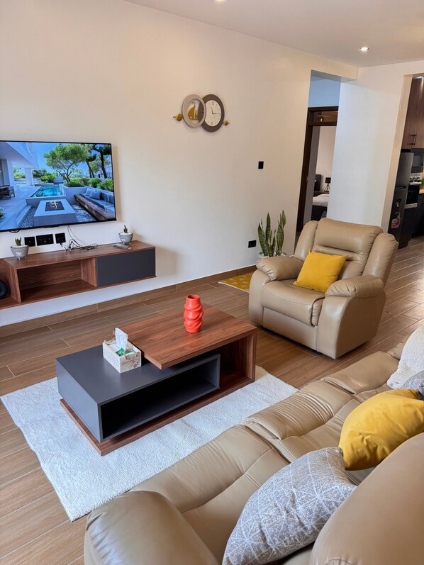 Un salon moderne doté d’un téléviseur à écran plat, d’un canapé en cuir, d’une table basse et d’un coin repas visible à travers une porte ouverte.
