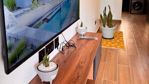 Un salon moderne avec un grand téléviseur à écran plat, une table basse en bois, des plantes en pot et un coin cuisine à l’arrière-plan.