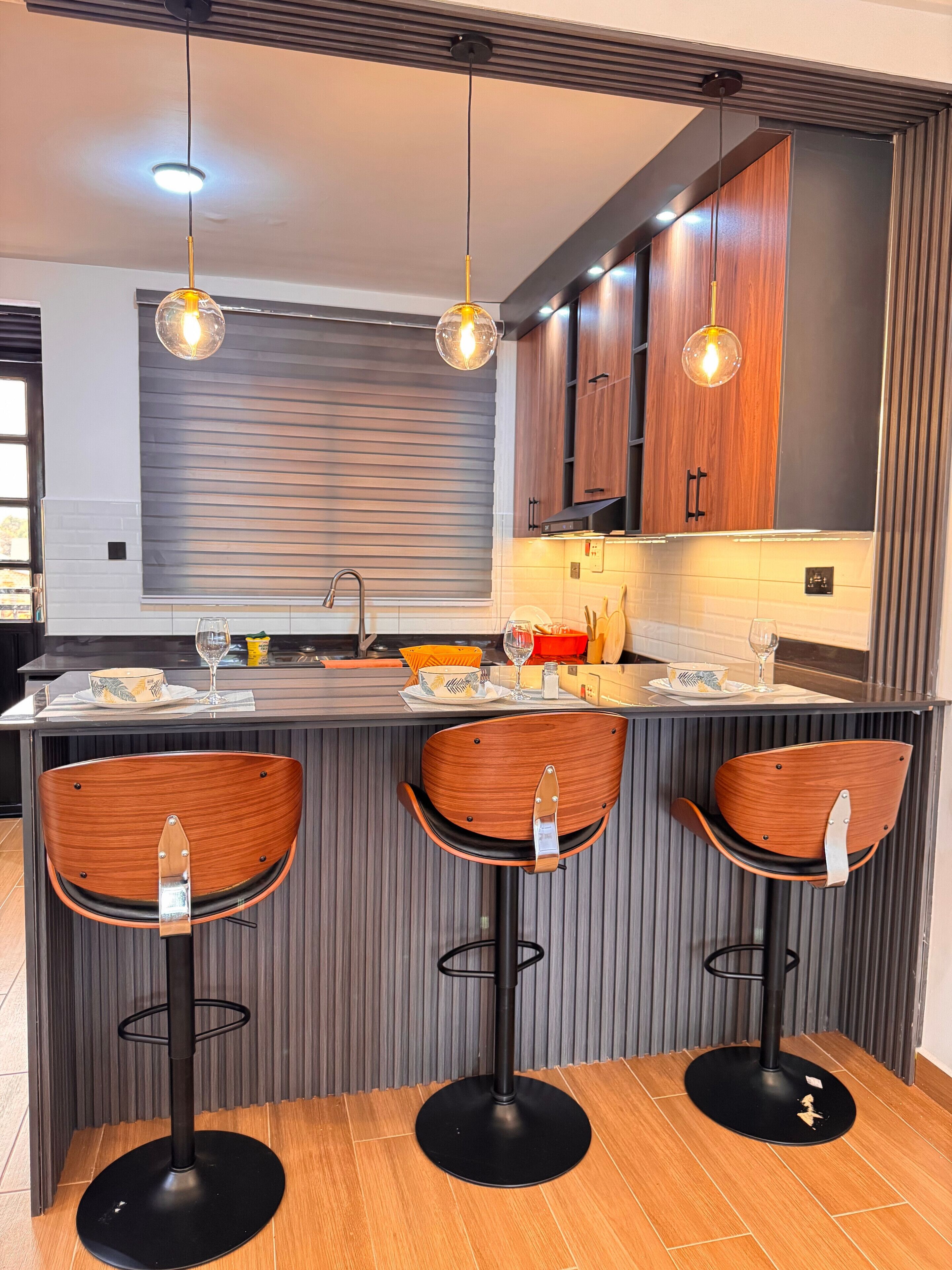 A modern kitchen with a central island, three wooden bar stools, pendant lighting, and wooden cabinets.