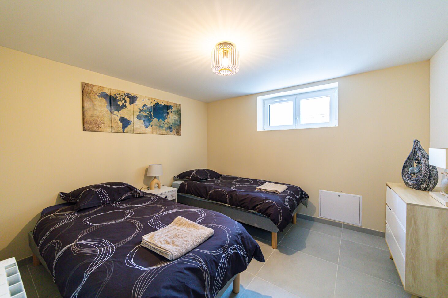 A bedroom with two beds, a map artwork, a window, and a desk with a lamp.