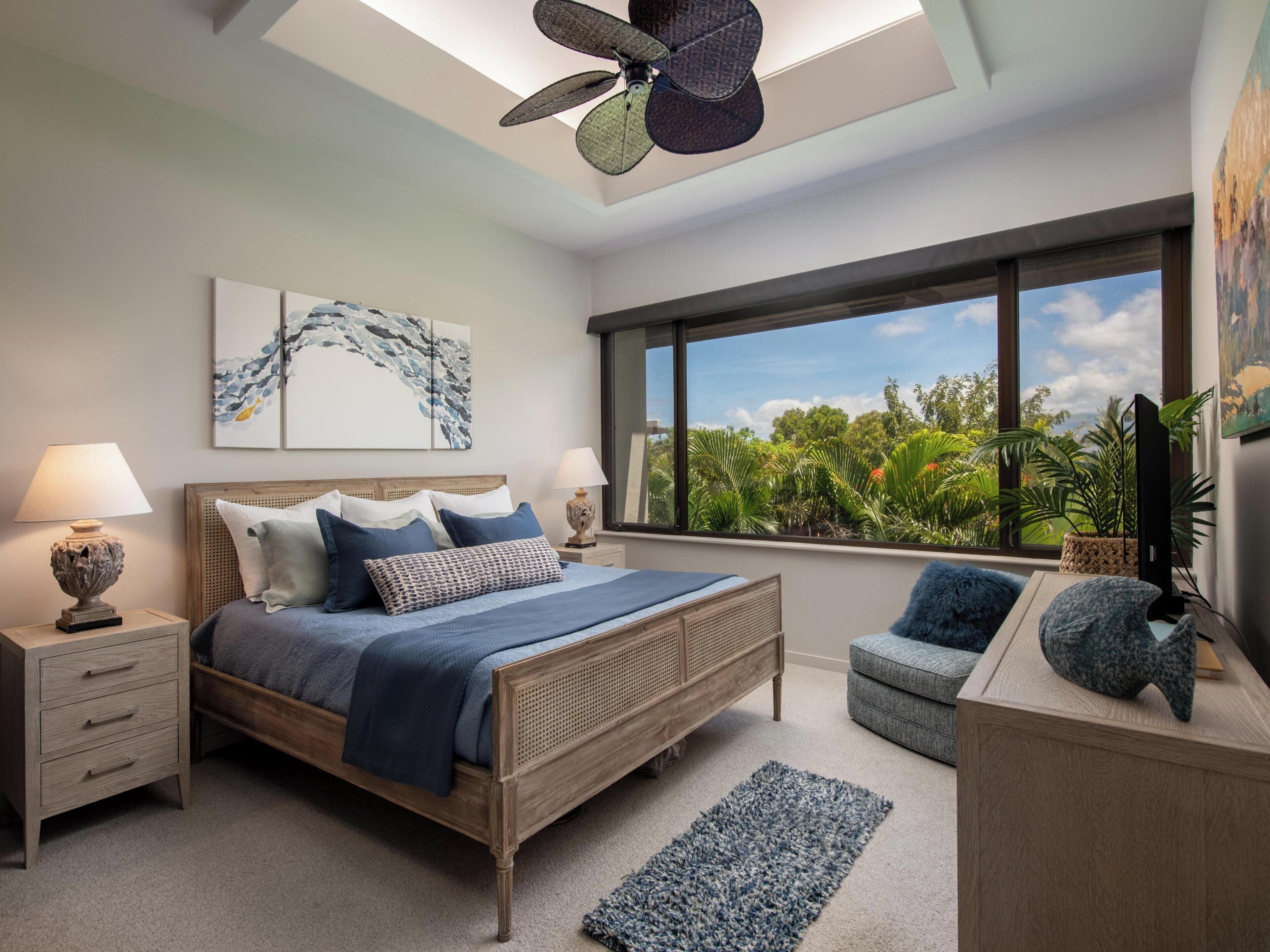 A bedroom with a bed, bedside tables, a ceiling fan, artwork, a large window with a view of greenery, and a desk with a computer.