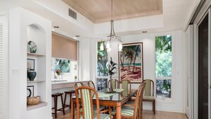 A dining area with a wooden table and chairs, a chandelier, and a view of greenery through large windows. - Great for Families & Professionally Managed | The Islands at Mauna Lani M1 (Kamuela)