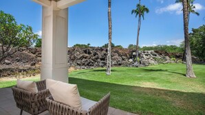A covered patio area with wicker furniture and a view of a landscaped lawn with palm trees and a rocky hillside. - Great for Families & Professionally Managed | The Islands at Mauna Lani M1 (Kamuela)