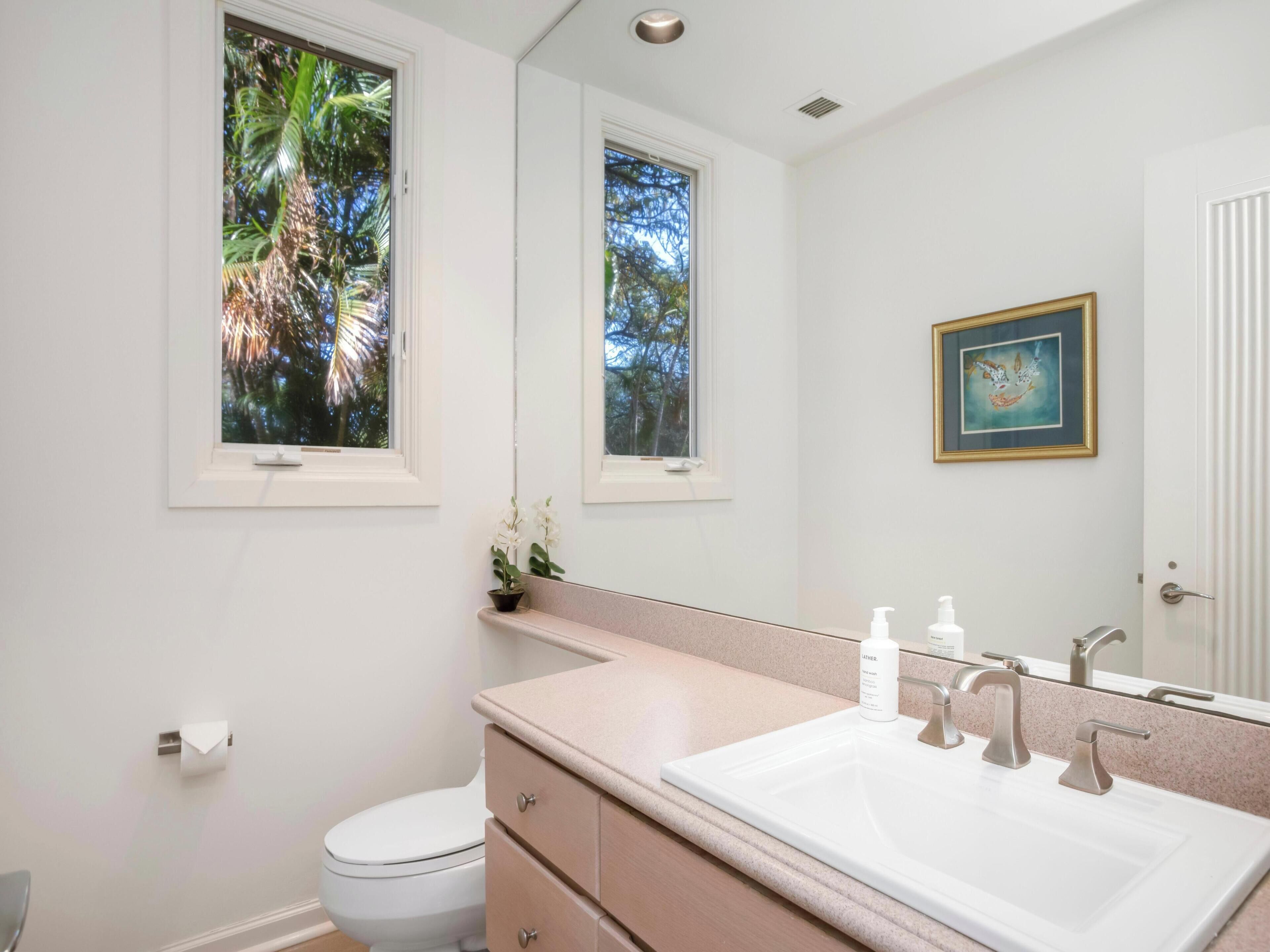 Ein Badezimmer mit einer weißen Toilette, einem Waschbecken mit Spiegel und einem Fenster mit Blick auf Palmen.