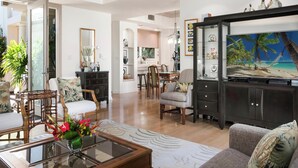 A spacious living room with a large flat-screen TV, comfortable seating, and a dining area visible through an open doorway. - Great for Families & Professionally Managed | The Islands at Mauna Lani M1 (Kamuela)