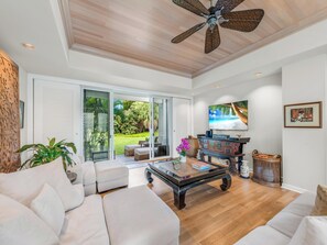A spacious living room with a ceiling fan, a large TV, and a view of a lush garden through sliding glass doors.