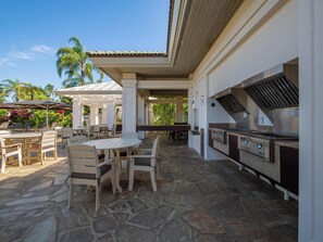 Une terrasse extérieure avec un sol en pierre, une table ronde et des chaises. On y trouve également un espace barbecue équipé d’une hotte en acier inoxydable.