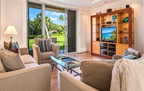 A living room with a glass coffee table, sofas, a TV on a wooden stand, and a sliding glass door leading to a garden.