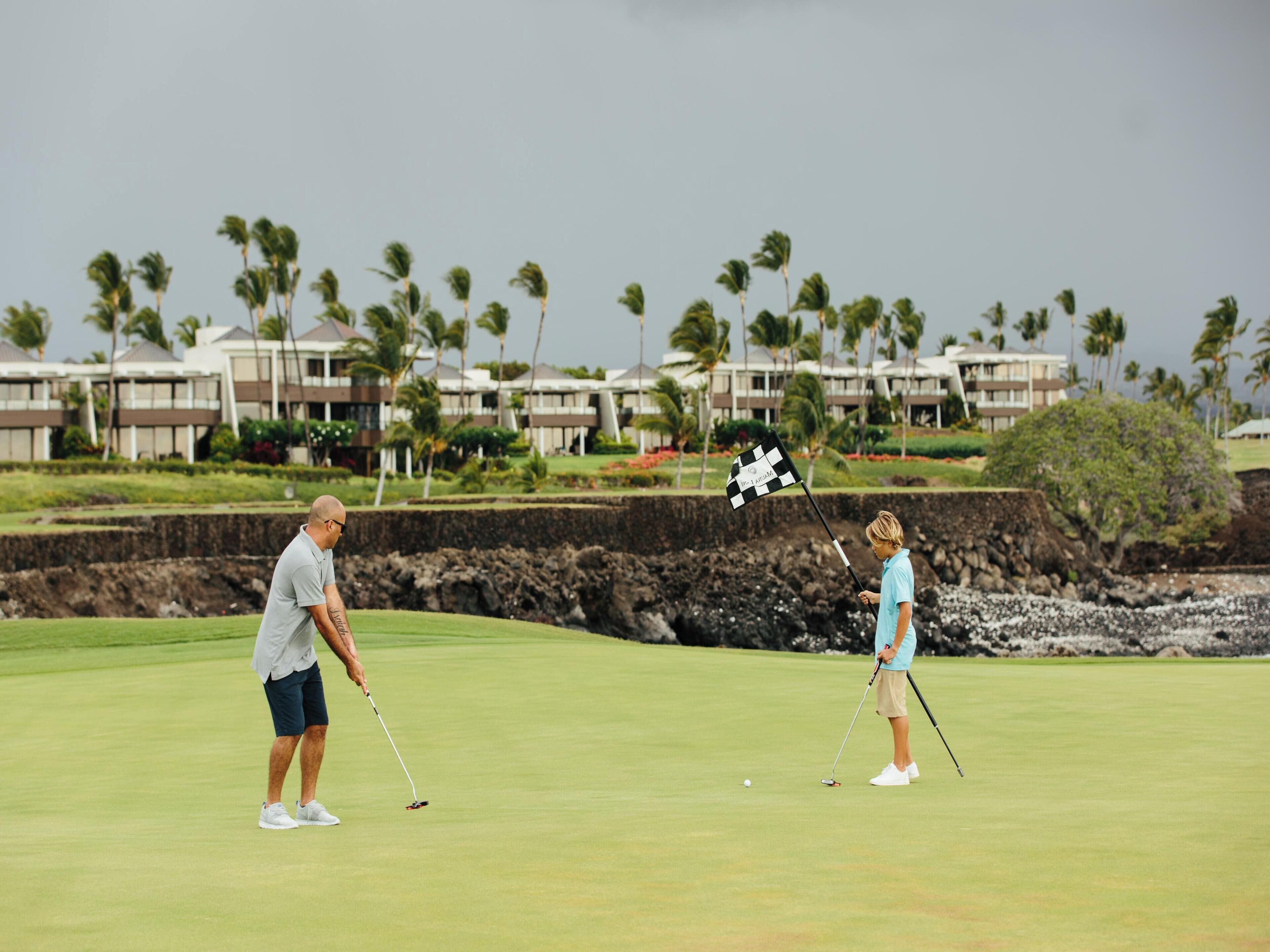 A golf course with two players, a flagstick, and a backdrop of palm trees and buildings.