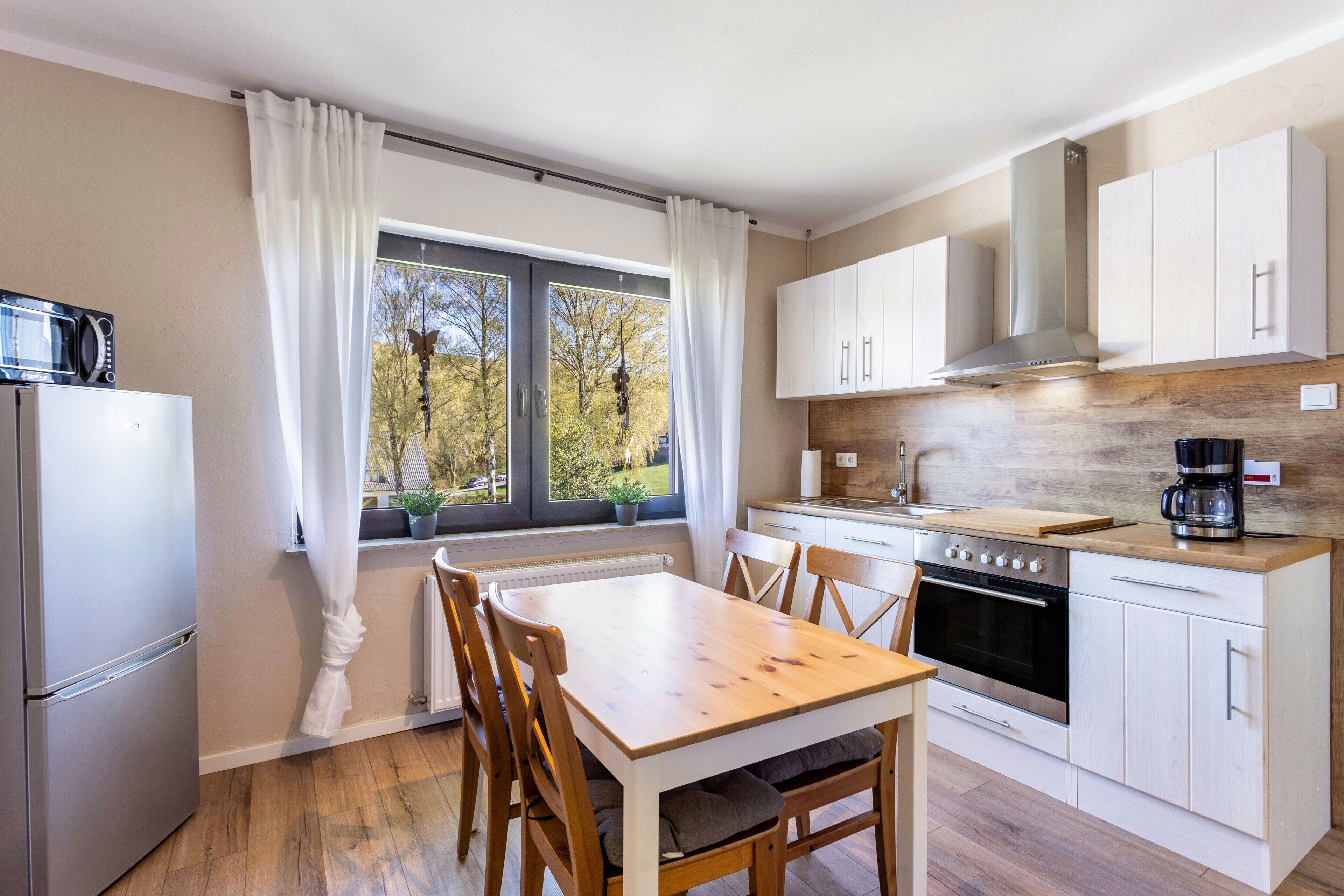 A modern kitchen with a dining table, chairs, a refrigerator, oven, cabinets, and a window with curtains.