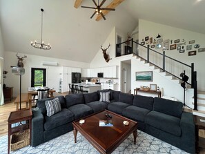 A spacious living room with a large sectional sofa, a wooden coffee table, and a dining area with a chandelier. - Stunning 5-bedroom house with AC in Franklin Perfect for visiting Downeast Maine (Franklin)