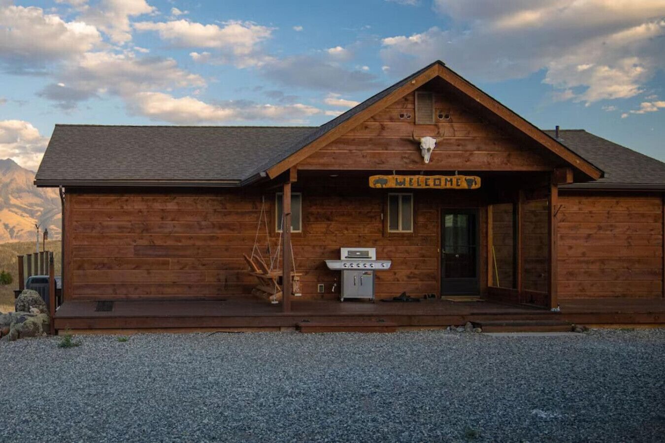 A wooden lodge with a 'Welcome' sign, a grill, and a mountain backdrop.